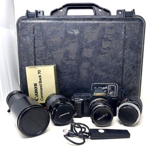 May include: A black Canon T70 camera body with a lens attached, two additional lenses, a Canon Command Back 70 box, a black remote shutter release cable, and a large black hard-shell carrying case with a handle and a Pelican brand sticker.