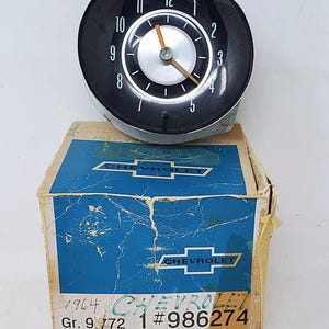 May include: A vintage black Chevrolet clock with white numerals and silver accents, resting on a blue and tan box. The clock face shows the time. The box has the Chevrolet logo and text that reads "1964 Chevrolet".