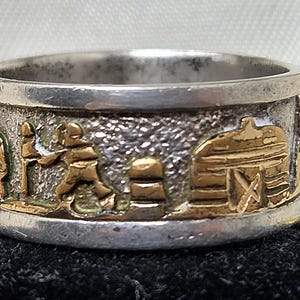 May include: A wide band ring featuring a textured silver-tone background with raised gold-tone figures depicting a person walking with a dog, a small pillar, and a barn structure. The ring has smooth, polished silver-tone edges at the top and bottom.