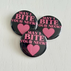 May include: Three black round buttons with the text "MAY I BITE YOUR NECK?" in pink, along with a pink heart. The buttons are arranged in a cluster on a white surface.