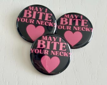 May include: Three black round buttons with the text "MAY I BITE YOUR NECK?" in pink, along with a pink heart. The buttons are arranged in a cluster on a white surface.