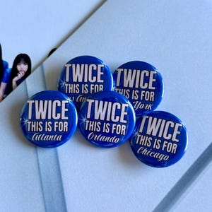 May include: A collection of five blue round buttons. Each button has the word "TWICE" in white, with the text "THIS IS FOR" and a city name. The cities are Atlanta, Orlando, Chicago, New York, and an additional one.
