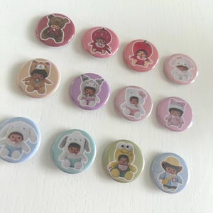 May include: A collection of round, colorful pinback buttons featuring cartoon characters in various animal costumes. The buttons are primarily pink, blue, and green, with detailed illustrations of bears, strawberries, and other cute designs. The buttons are arranged on a white surface.