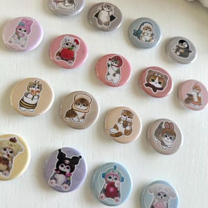 May include: A collection of round, colorful cat-themed button pins. Each pin features a different cartoon cat design, dressed in various outfits and accessories. The pins are in shades of pink, beige, gray, and blue, with a white background.
