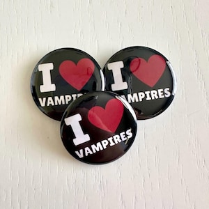 May include: Three black round buttons with the text "I <3 VAMPIRES" in white and a red heart. The buttons are glossy and have a pin back. The buttons are on a white wooden surface.