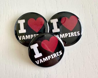 Puede incluir: Tres botones redondos negros con el texto "I <3 VAMPIRES" en blanco y un corazón rojo. Los botones son brillantes y tienen un alfiler en la parte posterior. Los botones están sobre una superficie de madera blanca.