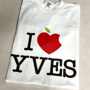May include: White t-shirt with a red apple graphic and the text "I YVES" in black. The apple has a bite taken out of it and a green leaf. The shirt is made of a soft material.
