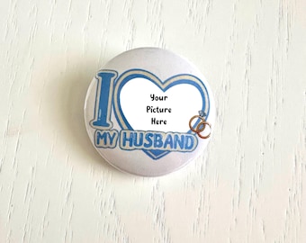 May include: A white button with a blue heart design and the text "I MY HUSBAND". The center of the heart is blank and says "Your Picture Here". Two gold wedding rings are on the right side of the heart.