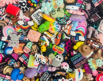 May include: A large collection of colourful, small, rubber charms with various designs and phrases. The charms feature animals, rainbows, hearts, and other cute designs. Some of the charms have text on them, such as "It's fine, I'm fine, everything's fine" and "Be the change".