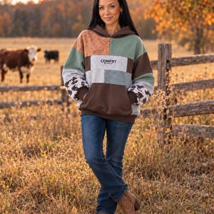 May include: A long-sleeve hooded sweatshirt featuring a patchwork design in shades of brown, sage green, and gray. The sleeves have a black and white cow print pattern. The center chest displays the text COMFRT. It is paired with blue denim jeans.