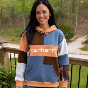 Peut inclure: Un sweat à capuche color-block composé de panneaux orange, marron, bleu et blanc. Le panneau central avant est orange avec le texte blanc "carhartt" imprimé. Le vêtement comprend une poche kangourou sur le devant et des cordons de serrage.