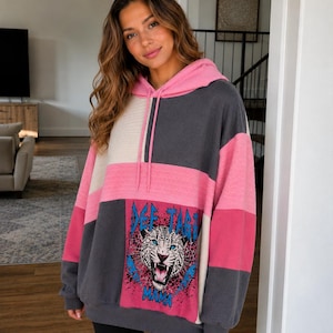 May include: Color-block hoodie in pink, gray, and white with a drawstring hood. The front features a graphic of a roaring tiger with the text "DEE TAH MAMA" in blue. The hoodie is worn with black leggings.