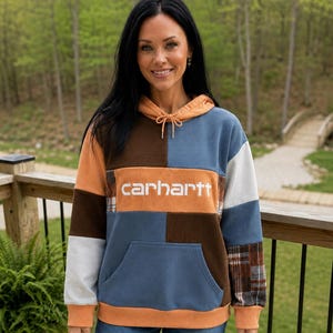 May include: A color-blocked corduroy hoodie featuring a patchwork design in shades of orange, brown, blue, and white. The garment includes a drawstring hood, a front kangaroo pocket, and the word carhartt printed in white across the chest.