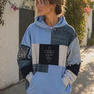 May include: A long-sleeve hooded sweatshirt featuring a patchwork design of light blue, dark blue denim, and grey fabric panels. The center front displays a black rectangular patch with a white cross graphic and the text God Has A Plan.
