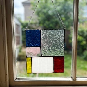 May include: A rectangular stained glass suncatcher featuring a grid of colored glass panels including deep blue, textured clear, pale pink, bright yellow, opaque white, and red, all held together by dark metal solder lines and suspended by a silver chain.