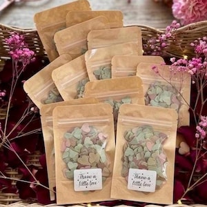 May include: Several small, clear plastic bags filled with heart-shaped confetti in pastel shades. The bags are sealed and arranged in a wicker basket, with pink flowers and rose petals. Each bag has a sticker that reads "Throw a little love."