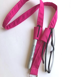 May include: A bright pink lanyard with a silver clasp, holding a black-framed pair of glasses and a gray Sharpie marker. The lanyard has a pocket for the marker and a loop to hold the glasses.