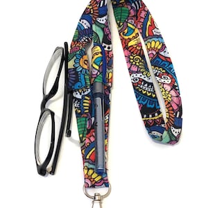 May include: A fabric lanyard featuring a vibrant, multicoloured floral and geometric pattern. The lanyard includes a built-in pen holder containing a blue Pilot pen, a silver-toned metal swivel lobster clasp at the bottom, and a pair of black-framed glasses.