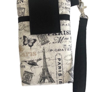 May include: A black and white fabric phone pouch with a Parisian theme. The pouch features a black strap, a black flap with a velcro closure, and a print of the Eiffel Tower, a butterfly, and a bird. The print also includes the words "Paris" and "Paris 18".