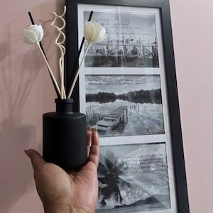 Puede incluir: Un difusor de aceites esenciales negro mate con cañas blancas en forma de flor. Un collage de fotos en blanco y negro enmarcado está en el fondo. El collage incluye imágenes de barcos, un muelle y palmeras.