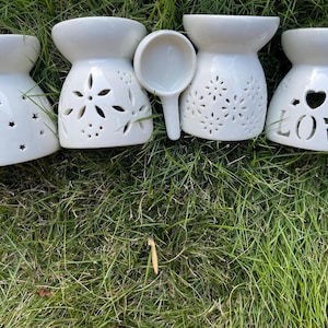 May include: Four white ceramic oil warmers with decorative cut-out designs, including stars, flowers, and hearts, and a small spoon. The warmers are arranged on a bed of green grass. The word "LOVE" is cut out on one warmer.
