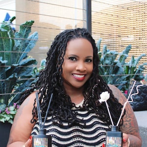 May include: Two black reed diffuser bottles with a decorative scene on the front, held by a person. The bottles have black reeds and a white flower and pink sticks. The person is wearing a black and white striped top and gold bracelets.