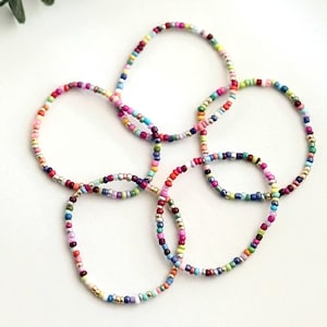 May include: A set of four colorful beaded bracelets with a mix of bright colors, including pink, blue, green, yellow, and purple. The bracelets are made with small, round beads and are strung on a thin, silver-colored thread.