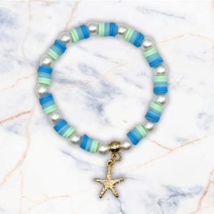 May include: A bracelet with blue and green disc beads, interspersed with white pearl beads. A gold-coloured starfish charm hangs from the centre. The bracelet is designed for a casual, beachy aesthetic. The bracelet's circumference is approximately 17 cm.