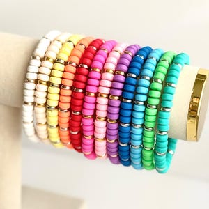 May include: A stack of colorful beaded bracelets in various shades of pink, orange, yellow, green, blue, and white. The bracelets are made of small, round beads and have a gold accent.