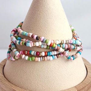 May include: A stack of colorful beaded bracelets. The bracelets are wrapped around a beige cone-shaped display. The beads are in various colors, including pink, blue, green, white, and gold. The bracelets are made of small, round beads.