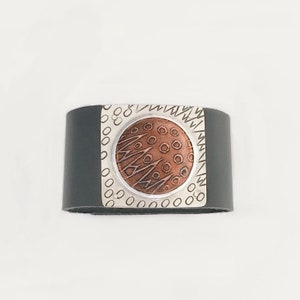 May include: A black leather cuff bracelet with a silver and copper circular design. The design features a textured copper circle with a silver border and a pattern of circles and lines.