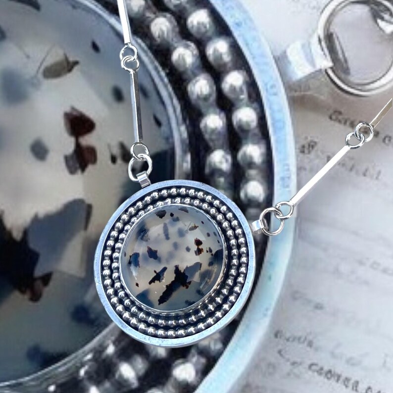 May include: A silver pendant necklace with a round, speckled stone center. The stone is encased in a silver bezel with a beaded border. The necklace chain is delicate and attached to the pendant with small silver bars.