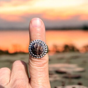 Puede incluir: Un anillo de plata con una piedra redonda y transparente que presenta un patr&oacute;n de &aacute;gata dendr&iacute;tica. La piedra est&aacute; engastada en un engaste de cuentas.