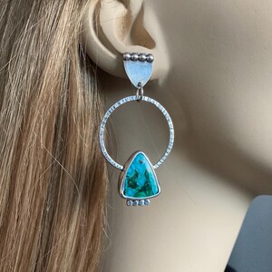 May include: Silver earrings with a geometric design. Each earring features a textured silver hoop, a triangular turquoise stone, and a small silver accent at the top and bottom. The earrings are designed to dangle and catch the light.