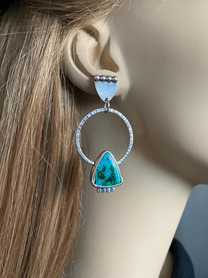 May include: Silver earrings with a geometric design. Each earring features a textured silver hoop, a triangular turquoise stone, and a small silver accent at the top and bottom. The earrings are designed to dangle and catch the light.