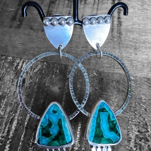 May include: A pair of silver earrings with a shield-shaped top and a large, textured hoop. The earrings feature a triangular turquoise and green stone. The earrings are displayed on a black stand against a gray and brown background.