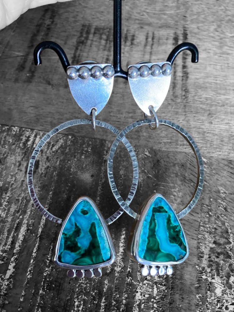 May include: A pair of silver earrings with a shield-shaped top and a large, textured hoop. The earrings feature a triangular turquoise and green stone. The earrings are displayed on a black stand against a gray and brown background.