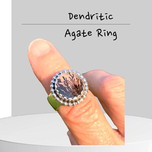 Puede incluir: Un anillo de plata con una piedra redonda de &aacute;gata dendr&iacute;tica engastada en un engaste de cuentas. El &aacute;gata tiene un fondo blanco transl&uacute;cido con patrones dendr&iacute;ticos negros.