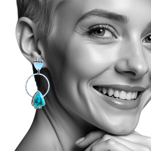 May include: Silver-toned dangle earrings with turquoise and light blue accents. The earrings feature a circular design with a turquoise triangle and a light blue, teardrop-shaped stone. The earrings are worn by a person with short blonde hair.