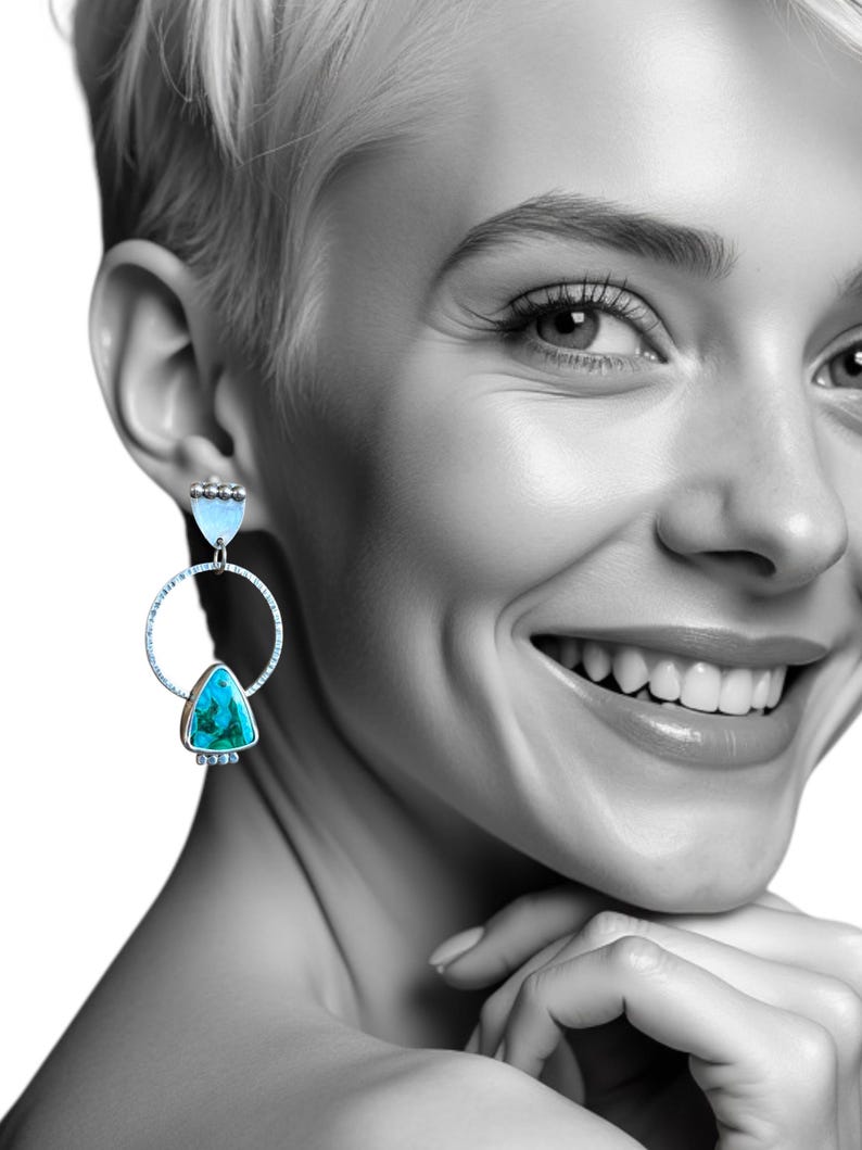 May include: Silver-toned dangle earrings with turquoise and light blue accents. The earrings feature a circular design with a turquoise triangle and a light blue, teardrop-shaped stone. The earrings are worn by a person with short blonde hair.