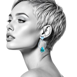 May include: A black and white image of a person wearing turquoise and silver earrings. The earrings feature a triangular turquoise stone suspended within a silver hoop. The person has short, light-colored hair and is looking to the side.