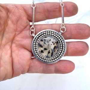 May include: A silver pendant necklace with a large, round, speckled stone center. The stone is surrounded by a beaded silver border and a smooth silver frame. The necklace has two silver bar accents connecting to the chain.
