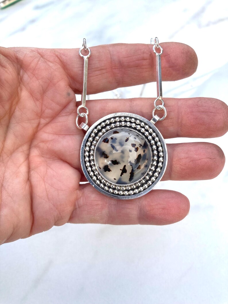 May include: A silver pendant necklace with a large, round, speckled stone center. The stone is surrounded by a beaded silver border and a smooth silver frame. The necklace has two silver bar accents connecting to the chain.