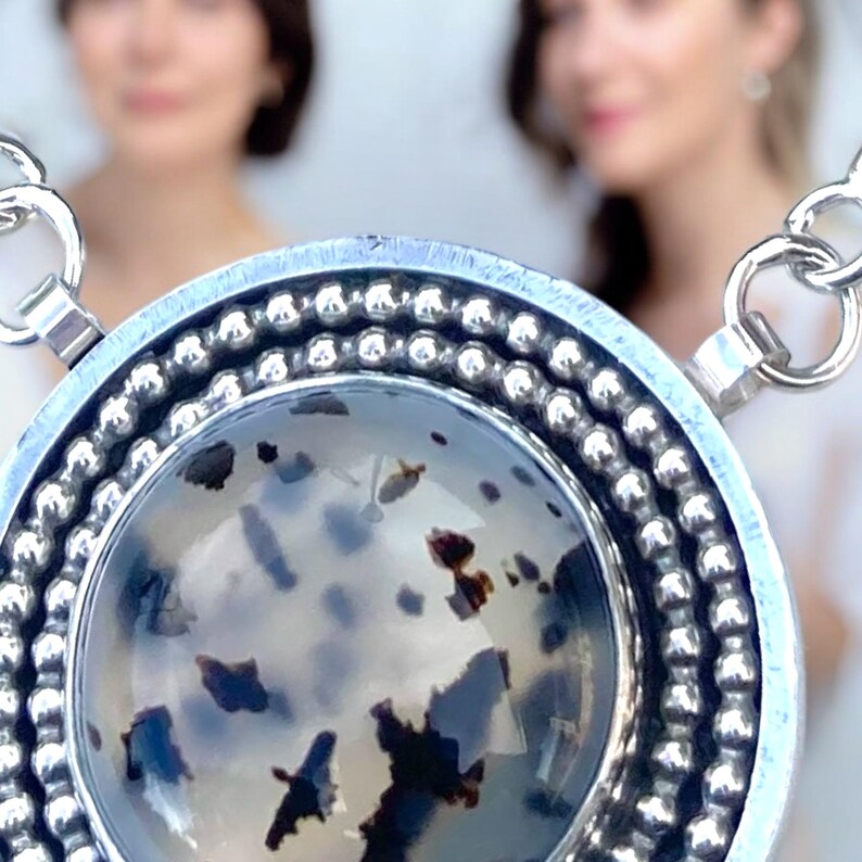 May include: A silver pendant necklace with a large, round, speckled stone. The stone is primarily white with dark brown and black inclusions. The pendant is surrounded by a beaded silver border and hangs from a silver chain.
