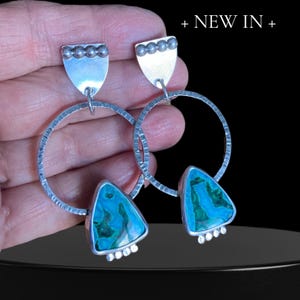 May include: Silver hoop earrings with turquoise and teal triangular stones. The earrings have a textured finish on the hoops and a small silver shield shape at the top. The text "+ NEW IN +" is at the top.