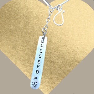 May include: A silver necklace with a rectangular pendant. The pendant is engraved with the word "BLESSED" in capital letters. Below the word is a heart symbol with the number .925. The necklace is displayed against a gold heart-shaped background.