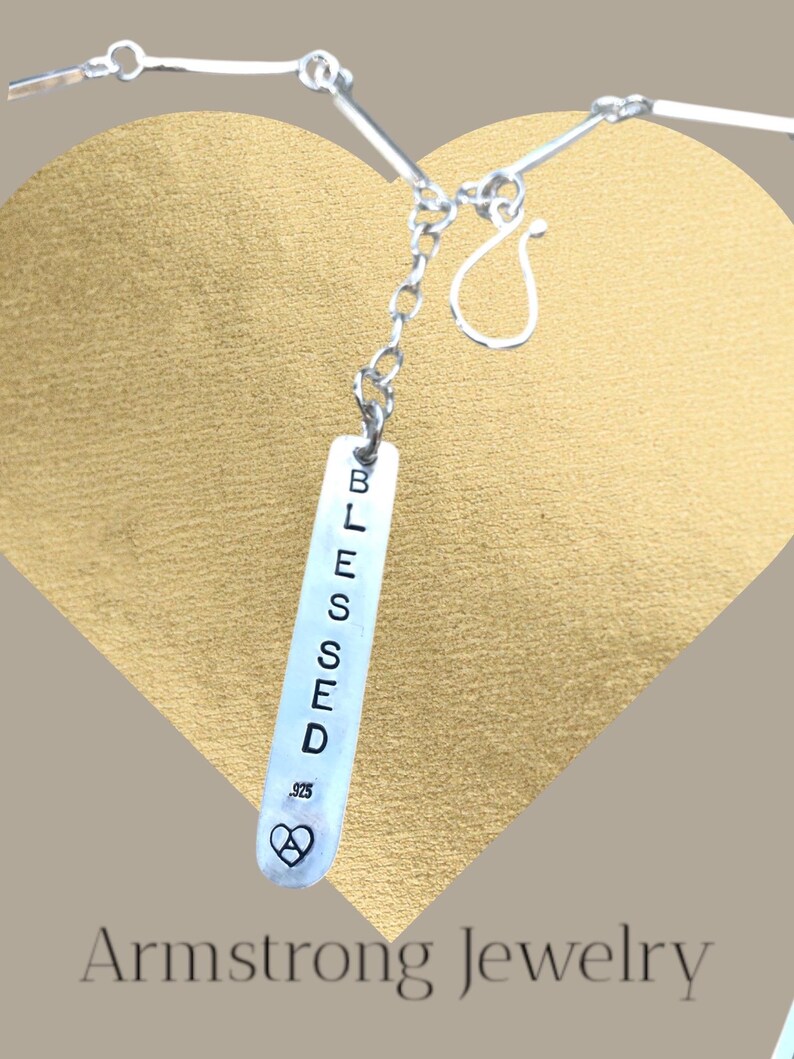 May include: A silver necklace with a rectangular pendant. The pendant is engraved with the word "BLESSED" in capital letters. Below the word is a heart symbol with the number .925. The necklace is displayed against a gold heart-shaped background.
