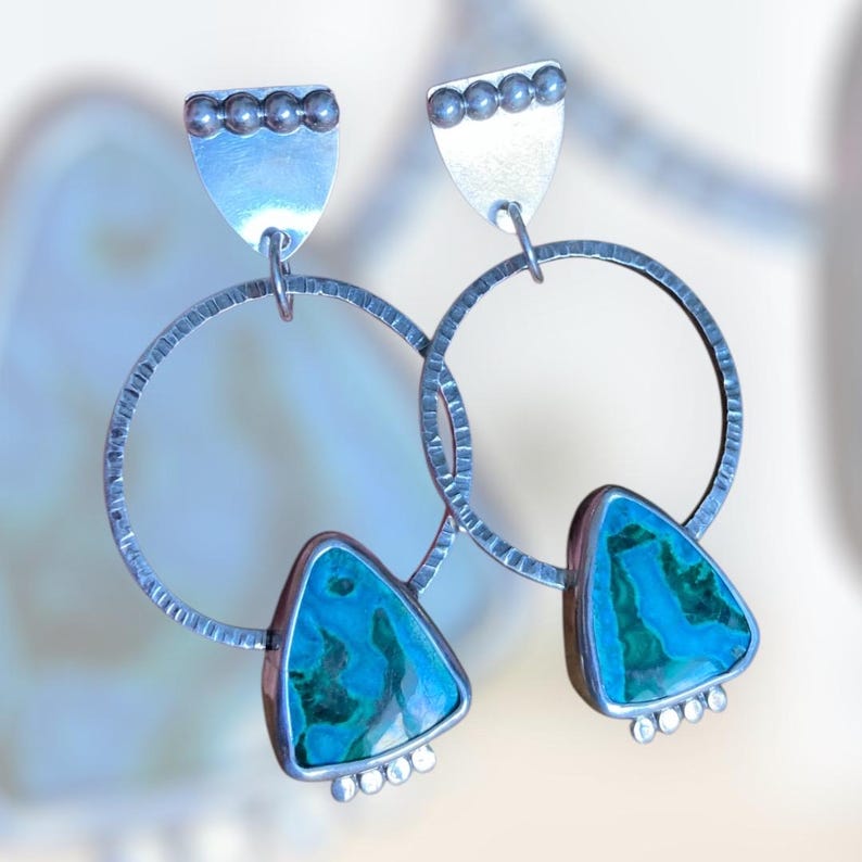 May include: A pair of silver earrings with a unique design. Each earring features a textured silver hoop, a triangular turquoise stone, and a small silver shield at the top. The turquoise stones have a blue and green marbled pattern.
