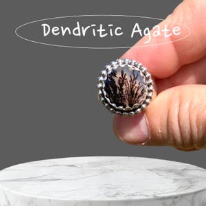 Puede incluir: Un anillo de plata con una piedra de &aacute;gata dendr&iacute;tica negra y blanca engastada en un engaste de cuentas. El anillo est&aacute; siendo sostenido por una mano.