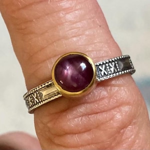 May include: A silver ring with a gold setting and a red ruby gemstone. The band is decorated with a repeating pattern.